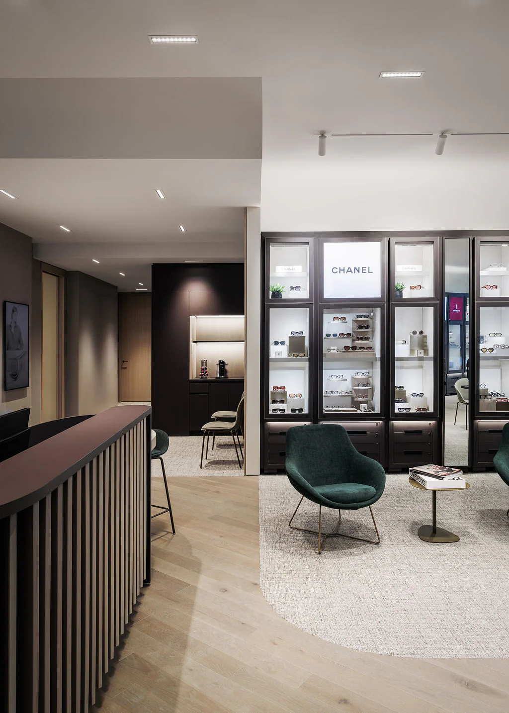 A modern, elegant waiting area with green velvet chairs, a small round table, and a curved dark wood reception desk. Behind glass shelves, Chanel-branded products are displayed. The space is well-lit and minimalist.