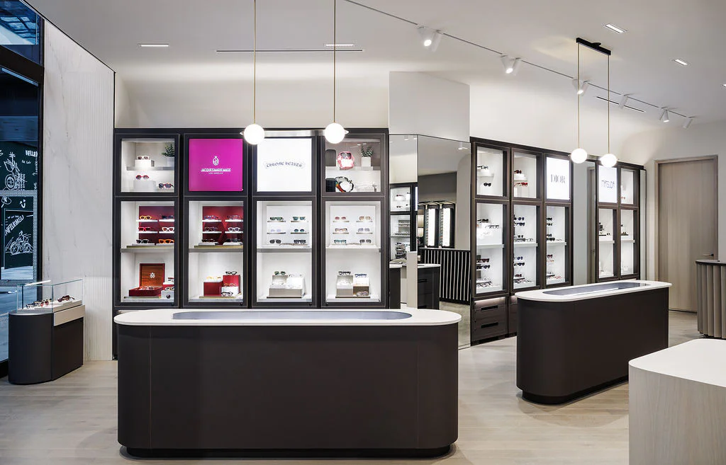 A modern eyewear store interior with sleek display cases showcasing various glasses and sunglasses. Two dark-colored counters stand in the foreground under pendant lights. The space is bright, organized, and minimalist.