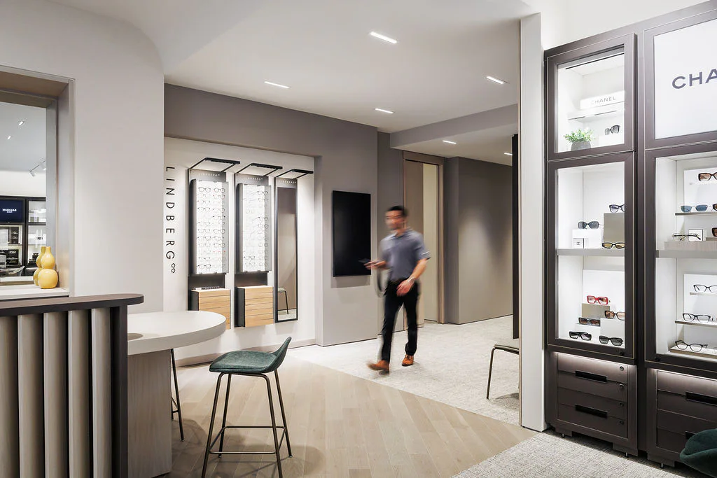 A modern eyewear store interior with sleek displays of glasses, neutral-colored walls and furniture, and a man walking through the space, appearing slightly blurred in motion.