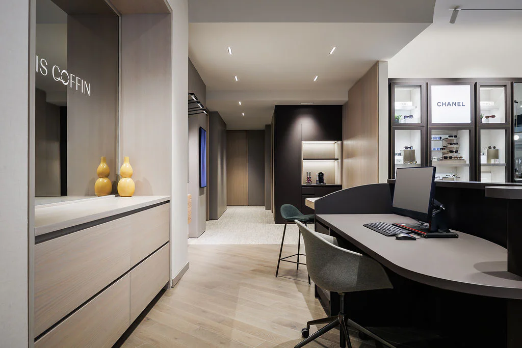 Modern optometry office with sleek wood flooring, a curved desk with a computer, gray chairs, display shelves with eyewear and CHANEL products, soft lighting, and decorative yellow vases on a side cabinet.
