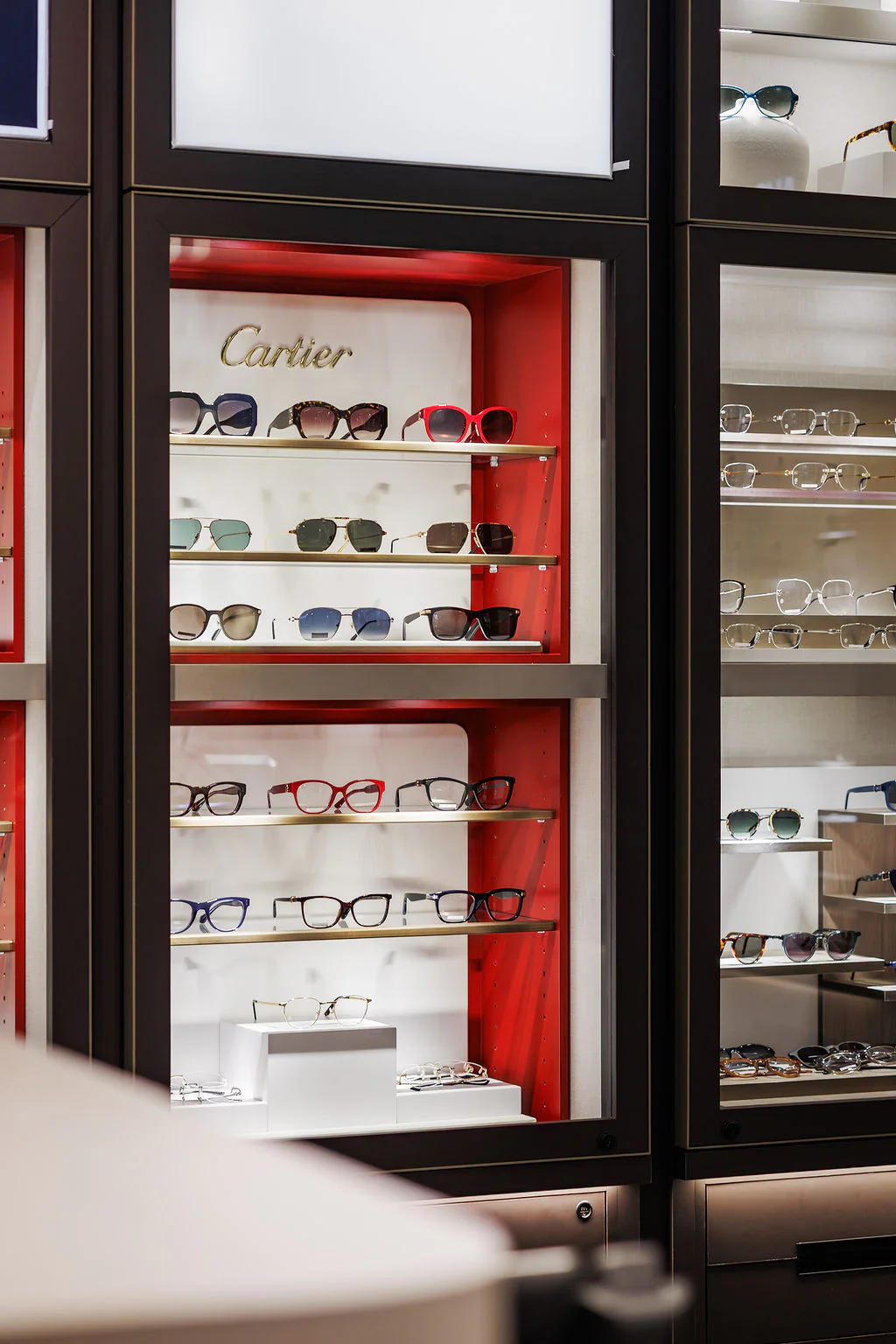 A display case featuring multiple rows of luxury Cartier eyeglasses and sunglasses, with a red and white background and a gold Cartier logo at the top. Adjacent showcases with other eyewear are partially visible.