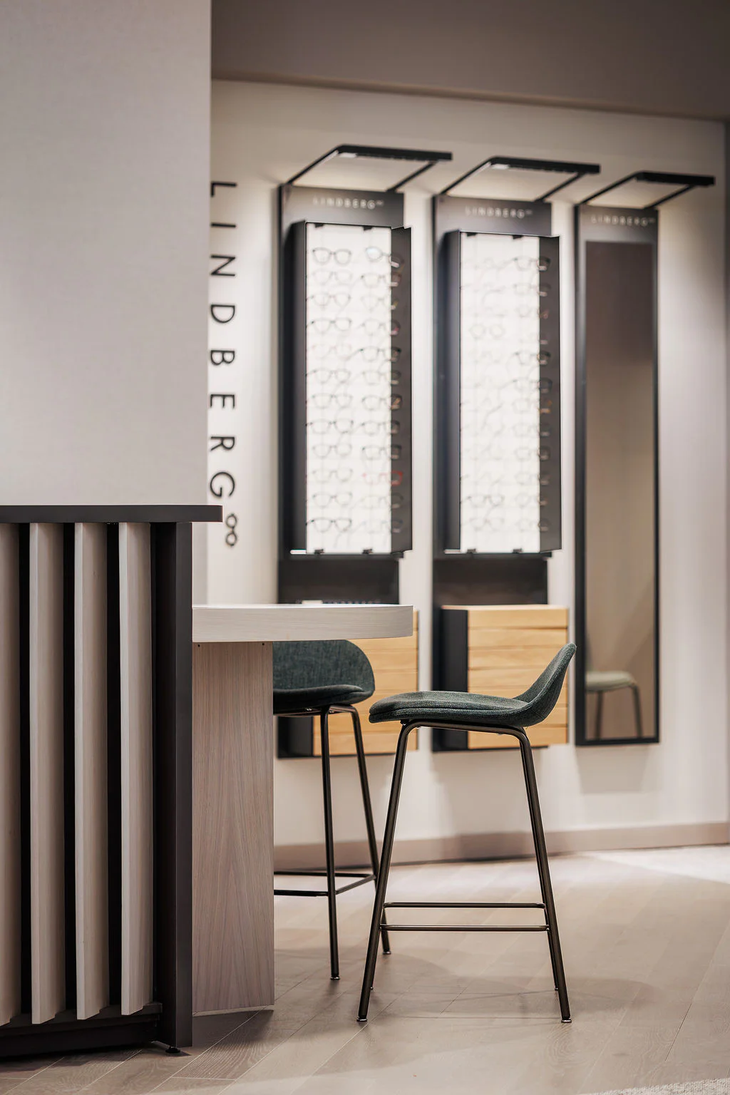Modern optician’s store interior with two dark green bar stools at a counter, and display racks of eyeglasses on the wall behind, labeled "Lindberg." The setting is clean, minimalistic, and well-lit.