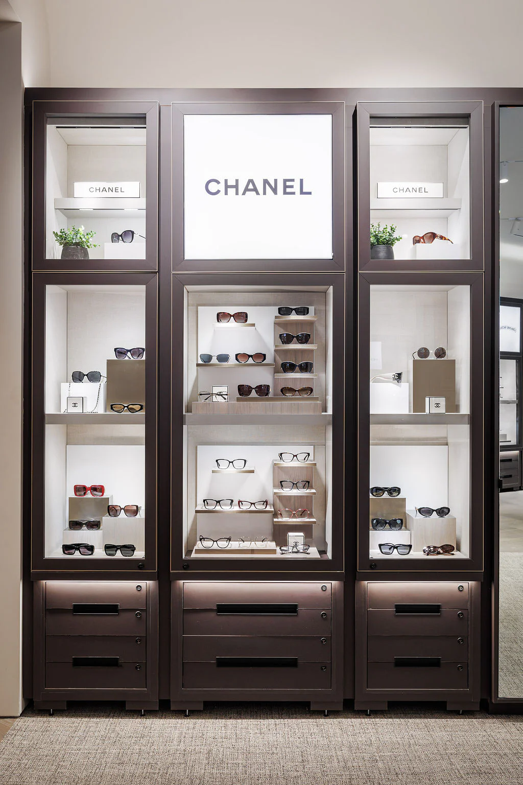 Display case with multiple shelves showcasing various sunglasses, with a prominent illuminated Chanel logo at the top center. Small potted plants decorate the corners, and the display has a modern, sleek design.