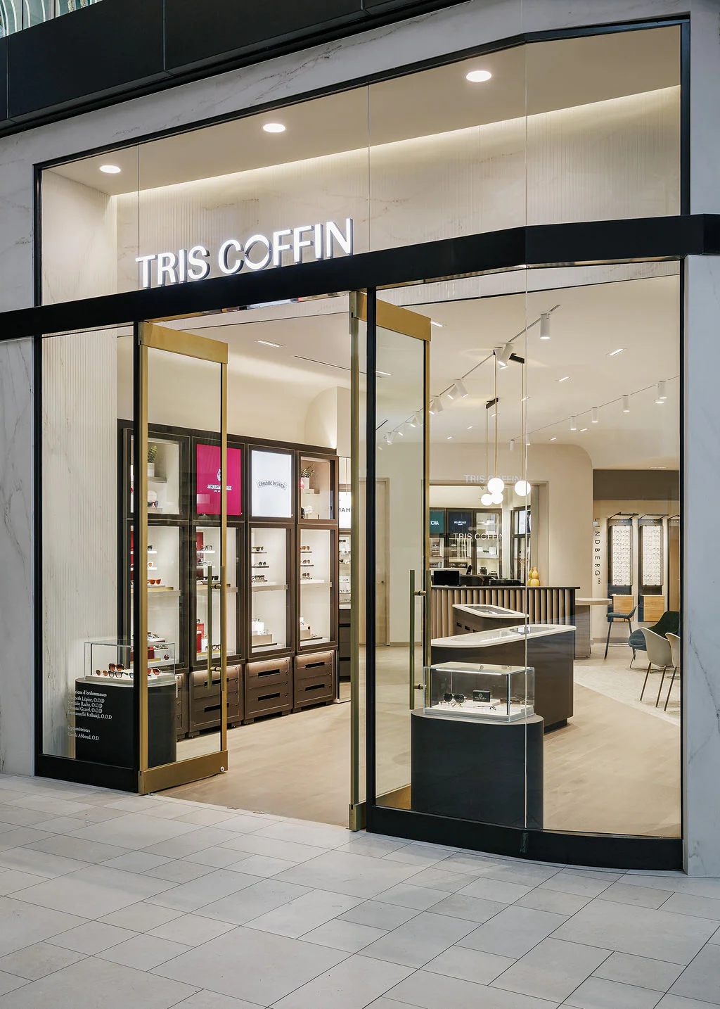 A modern eyewear store with glass doors open, displaying elegant eyewear displays, wooden counters, shelves, and bright lighting. The store sign above the entrance reads "TRIS COFFIN.