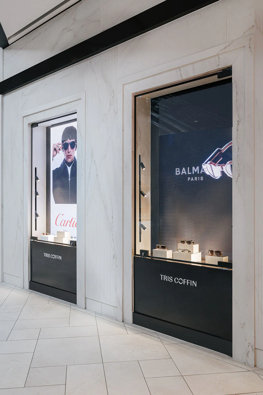 A storefront with two display windows; one has a Cartier ad featuring a man with sunglasses, the other showcases Balmain Paris products. The name "Tris Coffin" is on the bottom of both windows.