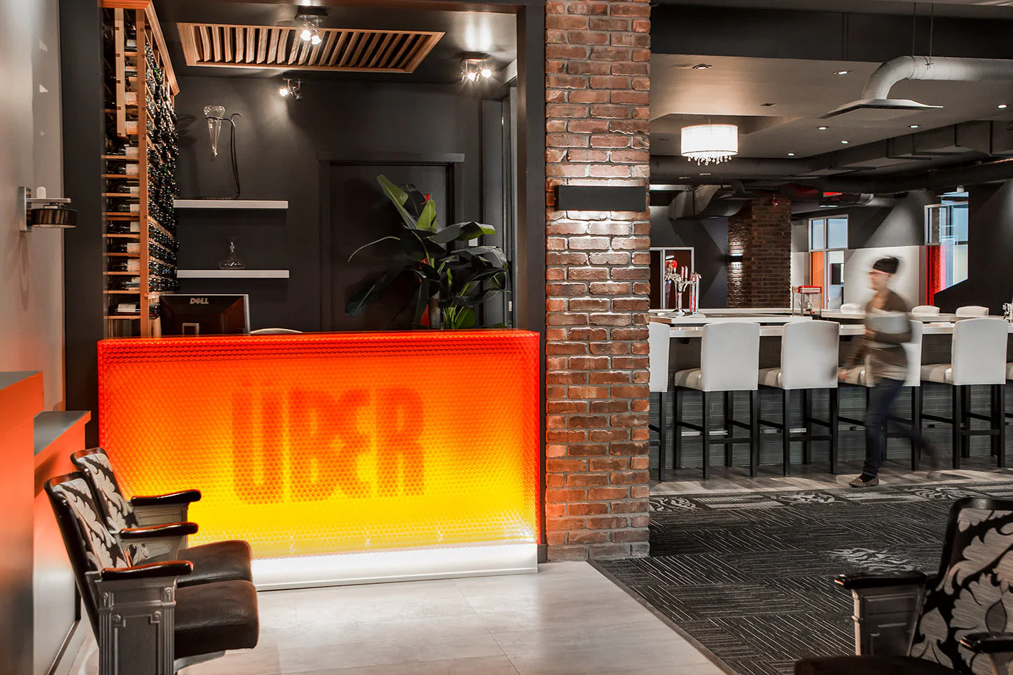 A modern office lobby with a glowing orange-yellow front desk labeled “UBER,” exposed brick walls, lounge chairs, and a blurred person walking past a white table and high chairs in the background.