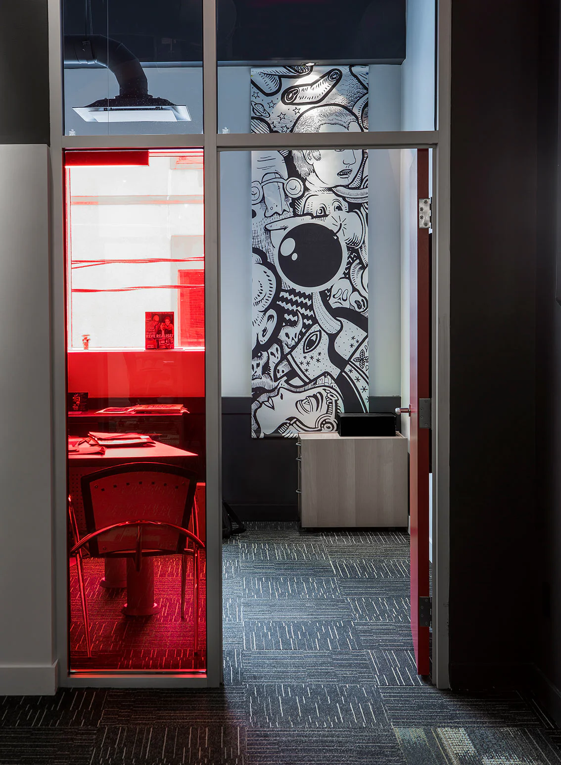 An office room viewed through a doorway, featuring a glass wall tinted red, modern furniture, and a large black-and-white mural with abstract faces and designs on the back wall.